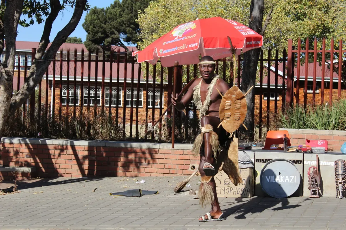 A performer working the pavement on Vilakazi Street, Soweto, in front of the precinct's trees and houses